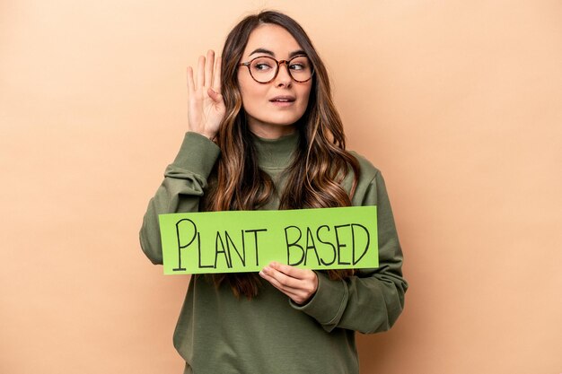Young caucasian woman holding a plant based placard isolated on beige background trying to listening a gossip