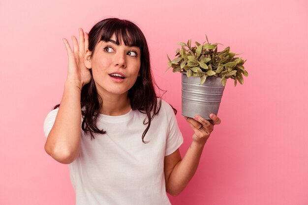 Young caucasian woman holding a plant isolated on pink background trying to listening a gossip.