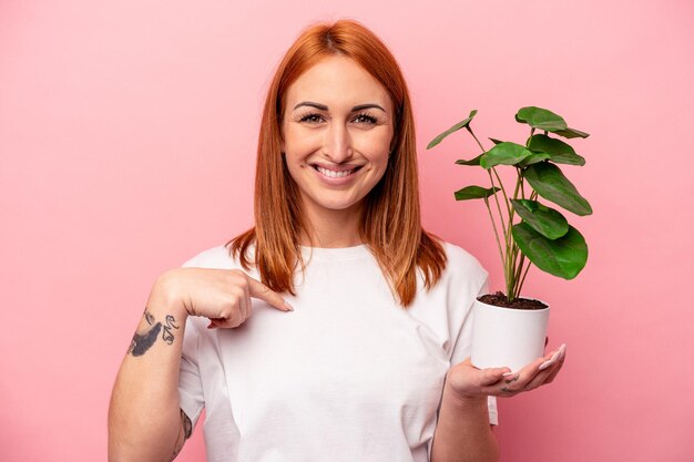 ピンクの背景に分離された植物を保持している若い白人女性ピンクの背景に分離された植物を保持している若い白人女性は、シャツのコピースペース、誇りと自信を持って手で指しています