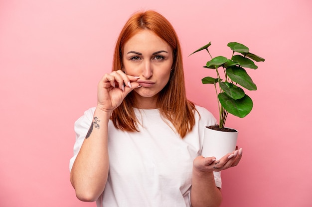 ピンクの背景に分離された植物を保持している若い白人女性秘密を保持している唇に指でピンクの背景に分離された植物を保持している若い白人女性。