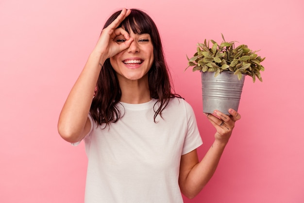 Young caucasian woman holding a plant isolated on pink wall excited keeping ok gesture on eye