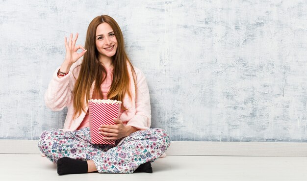 Young caucasian woman holding a popcorn bucket cheerful and confident showing ok gesture.