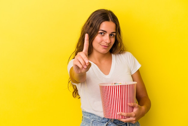 Young caucasian woman holding a popcorns isolated on yellow background  showing number one with finger.