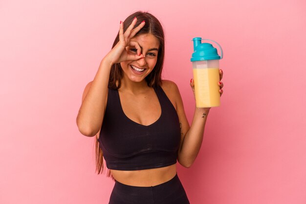 Young caucasian woman holding a protein shake isolated on yellow background excited keeping ok gesture on eye.