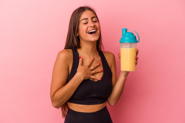 Young caucasian woman holding a protein shake isolated on yellow background laughs out loudly keeping hand on chest.