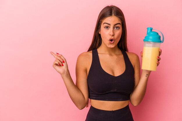 Young caucasian woman holding a protein shake isolated on yellow background pointing to the side