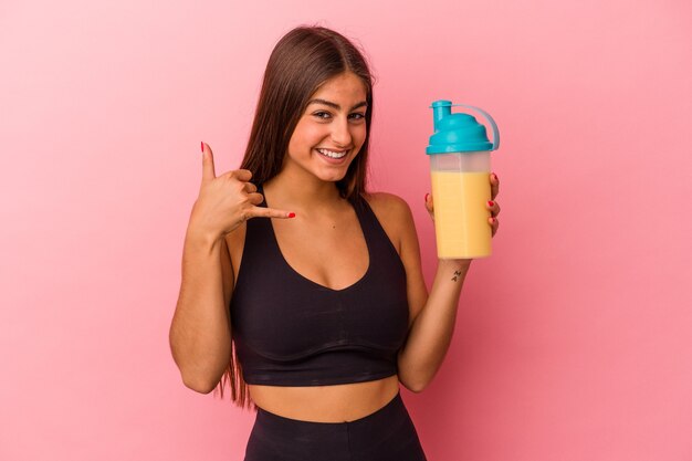 Young caucasian woman holding a protein shake isolated on yellow background showing a mobile phone call gesture with fingers.