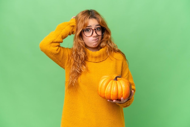 Young caucasian woman holding a pumpkin isolated on green screen chroma key background having doubts