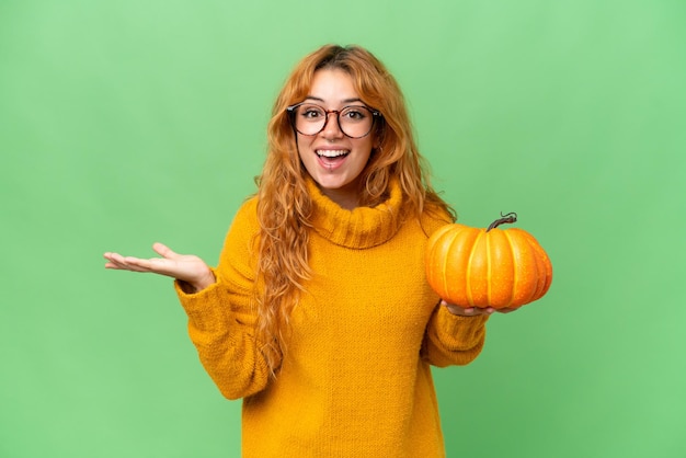 Young caucasian woman holding a pumpkin isolated on green screen chroma key background with shocked facial expression