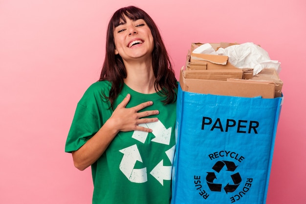 Young caucasian woman holding a recycled cardboard bag isolated on pink wall laughs out loudly keeping hand on chest.