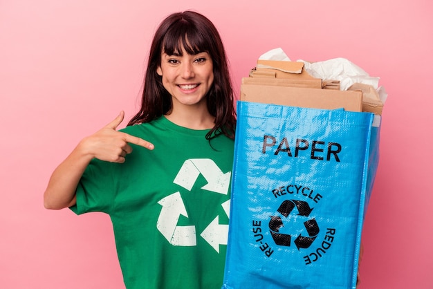 Young caucasian woman holding a recycled cardboard bag isolated on pink wall person pointing by hand to a shirt copy space, proud and confident