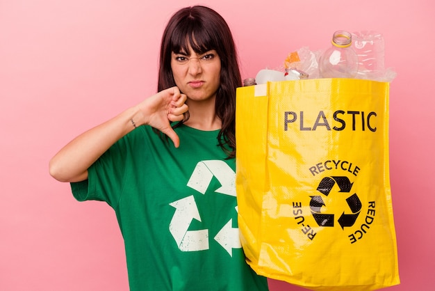 Young caucasian woman holding a recycled plastic bag isolated on pink background showing a dislike gesture, thumbs down. Disagreement concept.