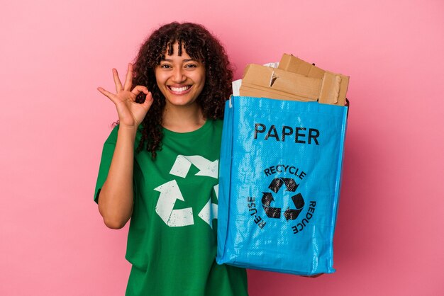 ピンクの背景に分離されたリサイクルプラスチックを保持している若い白人女性は、陽気で自信を持って大丈夫なジェスチャーを示しています。