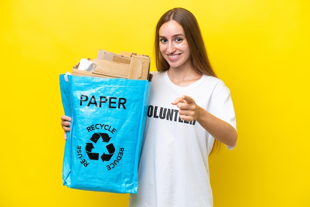 Young caucasian woman holding a recycling bag full of paper to recycle isolated on yellow background points finger at you with a confident expression