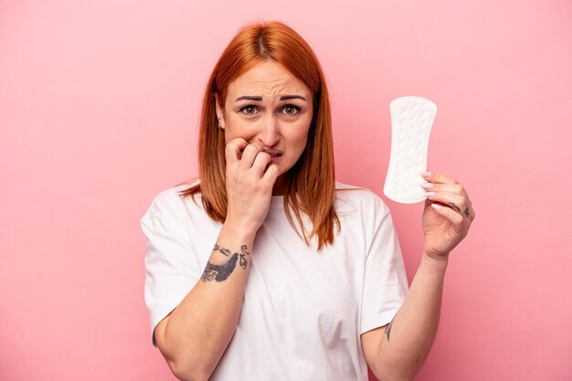 Foto giovane donna caucasica che tiene un assorbente sanitario isolato su sfondo rosa che si morde le unghie nervose e molto ansiose