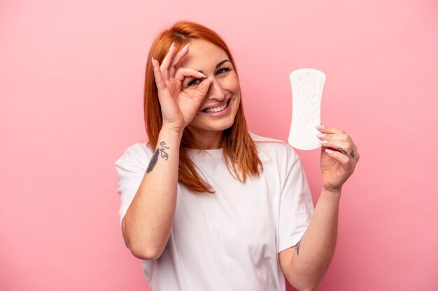 Young caucasian woman holding sanitary napkin isolated on pink background excited keeping ok gesture on eye