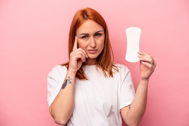 Young caucasian woman holding sanitary napkin isolated on pink background pointing temple with finger thinking focused on a task