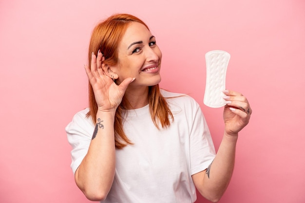 Young caucasian woman holding sanitary napkin isolated on pink background trying to listening a gossip