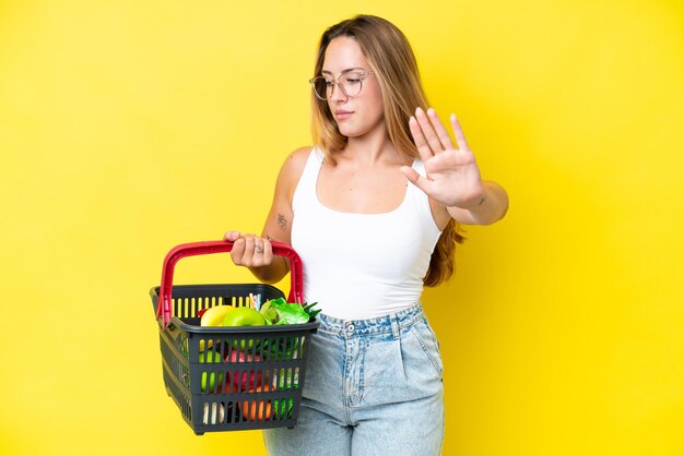 Foto giovane donna caucasica che tiene un carrello pieno di cibo isolato su sfondo giallo che fa gesto di arresto e deluso