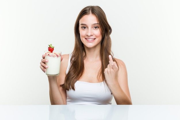 Young caucasian woman holding a smoothie pointing with finger at you as if inviting come closer.