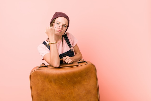 Young caucasian woman holding a suitcase showing fist to with aggressive facial expression.