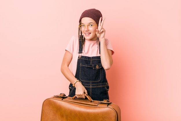 Young caucasian woman holding a suitcase showing victory sign and smiling broadly.