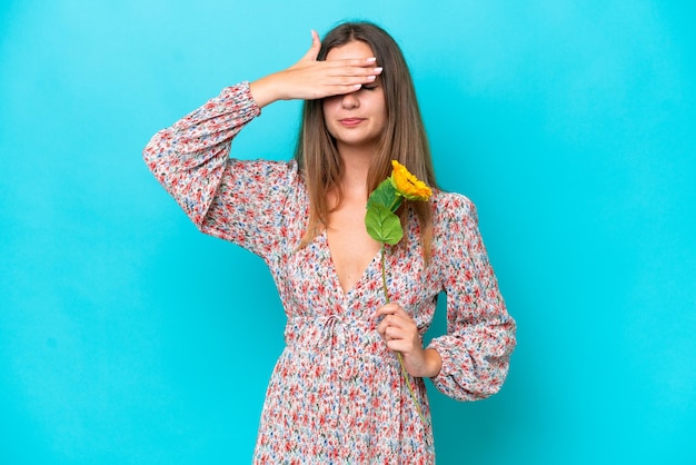 Young caucasian woman holding sunflower isolated on blue background covering eyes by hands Do not want to see something