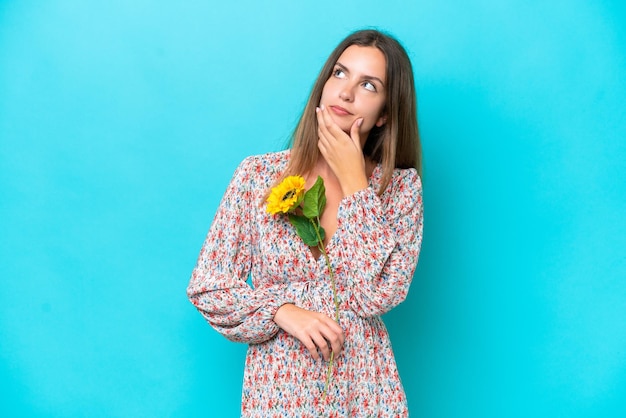 Giovane donna caucasica che tiene il girasole isolato su sfondo blu con dubbi