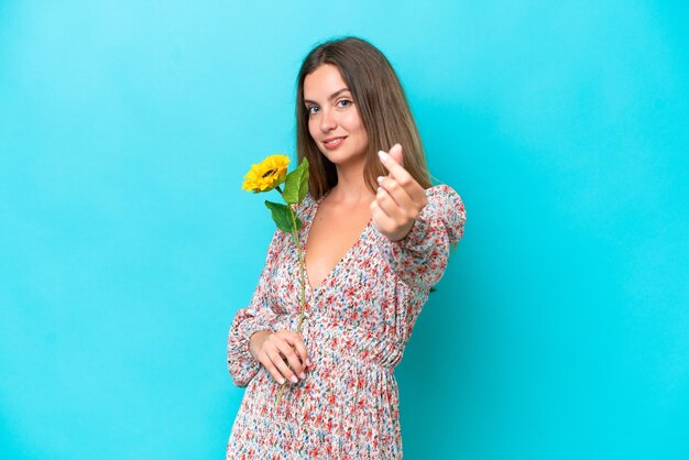 Young caucasian woman holding sunflower isolated on blue background making money gesture