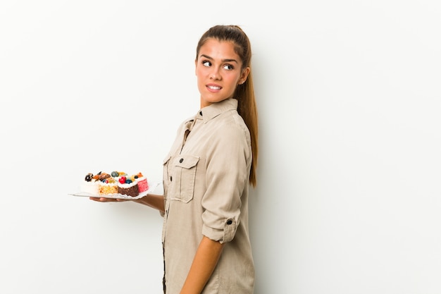 Young caucasian woman holding sweet cakes looks aside smiling, cheerful and pleasant.