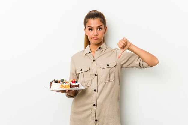 嫌いなジェスチャー、親指ダウンを示す甘いケーキを保持している若い白人女性。不一致の概念。