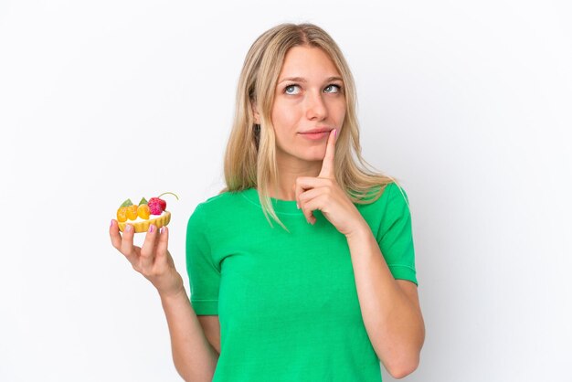 Young caucasian woman holding a tartlet isolated on white background having doubts while looking up