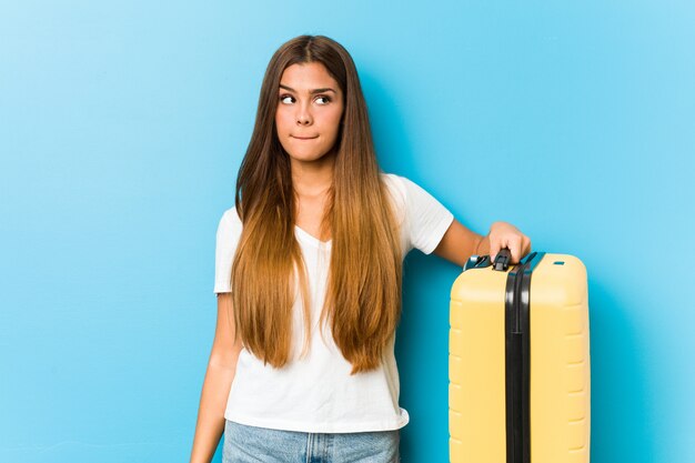 Young caucasian woman holding a travel suitcase confused, feels doubtful and unsure.