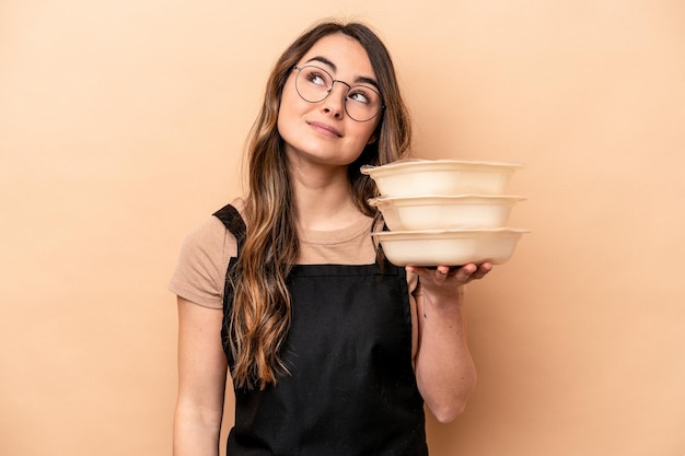 Молодая кавказская женщина, держащая посуду на бежевом фоне, мечтает о достижении целей и задач
