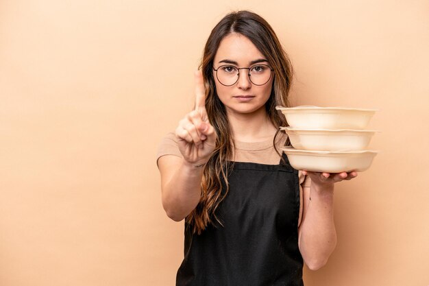 Young caucasian woman holding tupperware isolated on beige background showing number one with finger