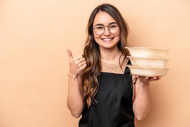 Молодая кавказская женщина, держащая посуду на бежевом фоне, улыбается и поднимает большой палец вверх