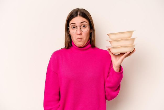 Young caucasian woman holding tupperware isolated on white background shrugs shoulders and open eyes confused