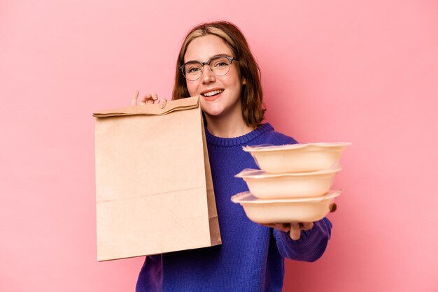 Young caucasian woman holding tupperware and takeaway bag isolated on pink background