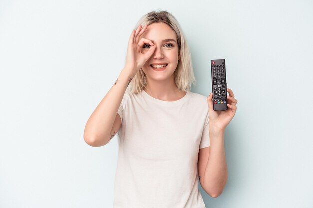 Молодая кавказская женщина, держащая контроллер телевизора, изолирована на синем фоне, взволнована, держа на глазах хорошо жест.