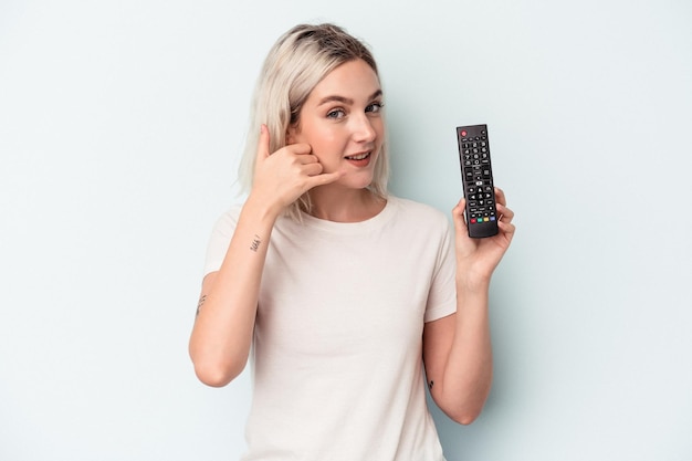 指で携帯電話の呼び出しジェスチャーを示す青い背景に分離されたテレビコントローラーを保持している若い白人女性。