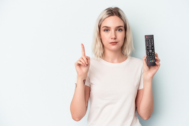 Young caucasian woman holding a tv controller isolated on blue background showing number one with finger.