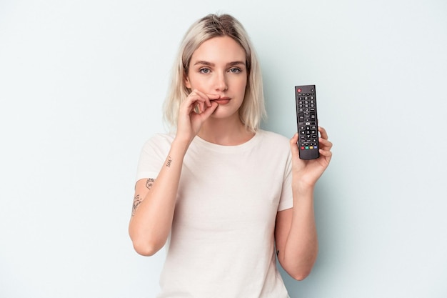 Young caucasian woman holding a tv controller isolated on blue background with fingers on lips keeping a secret.