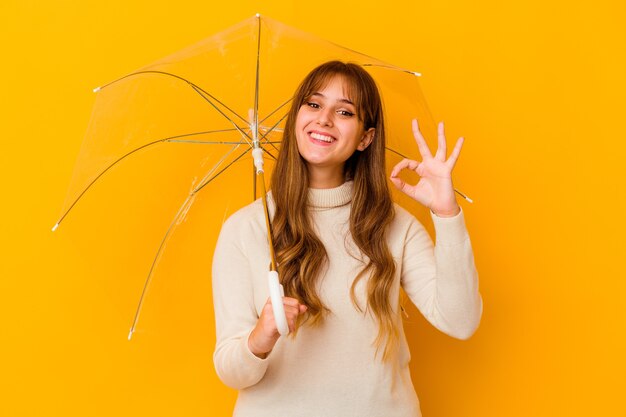 傘をさしている若い白人女性は、陽気で自信を持って大丈夫なジェスチャーを示しています。
