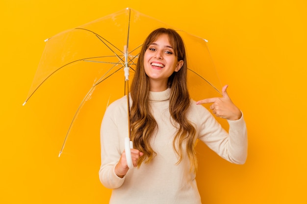 Young caucasian woman holding a umbrella isolated person pointing by hand to a shirt copy space, proud and confident