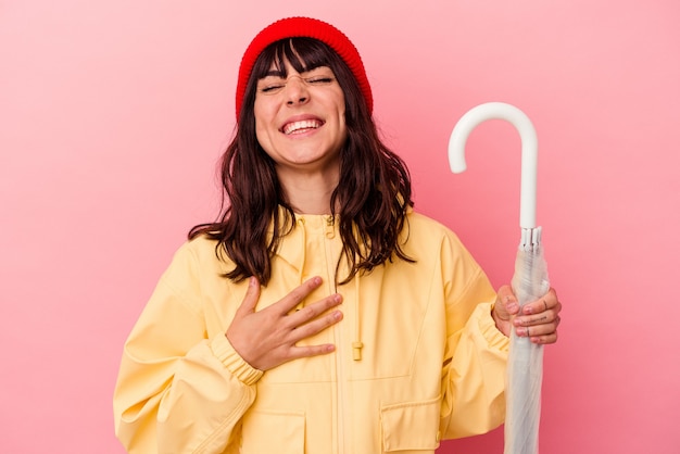 Young caucasian woman holding an umbrella isolated on pink background laughs out loudly keeping hand on chest.