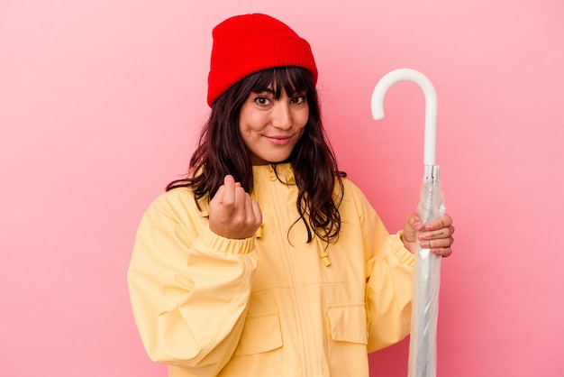 Young caucasian woman holding an umbrella isolated on pink background pointing with finger at you as if inviting come closer.