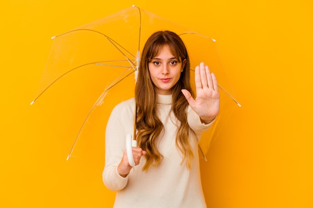 Young caucasian woman holding a umbrella isolated standing with outstretched hand showing stop sign, preventing you.