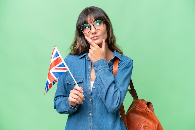 Young caucasian woman holding an United Kingdom flag over isolated background having doubts