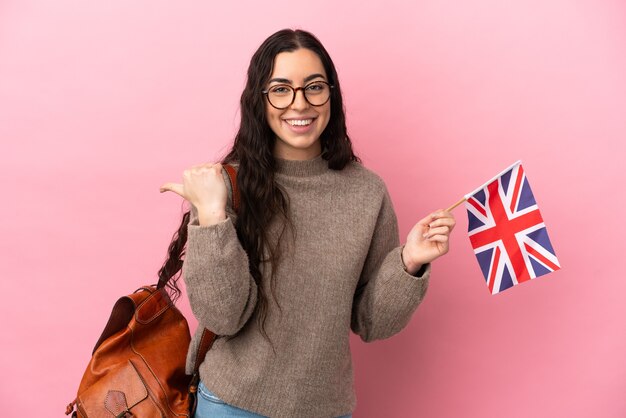 Young caucasian woman holding an United Kingdom flag isolated on pink background pointing to the side to present a product