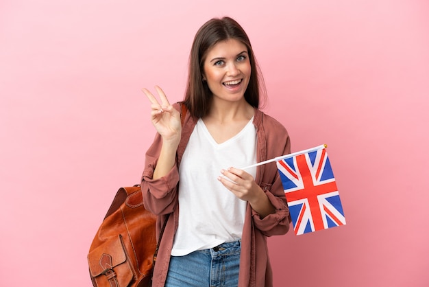 Young caucasian woman holding an United Kingdom flag isolated on pink background smiling and showing victory sign